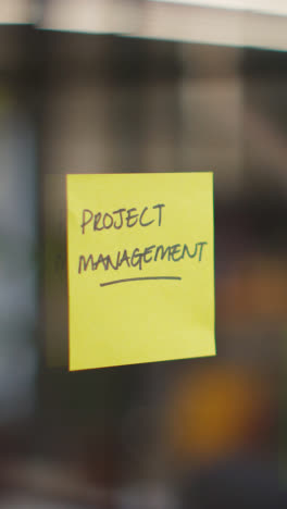 Vertical-Video-Close-Up-Of-Woman-Putting-Sticky-Note-With-Project-Management-Written-On-It-Onto-Transparent-Screen-In-Office-